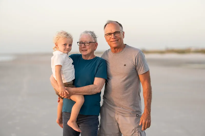 Three generations pose together on the beach—a child, two adults, and an older adult in neutral tones against the calm water.