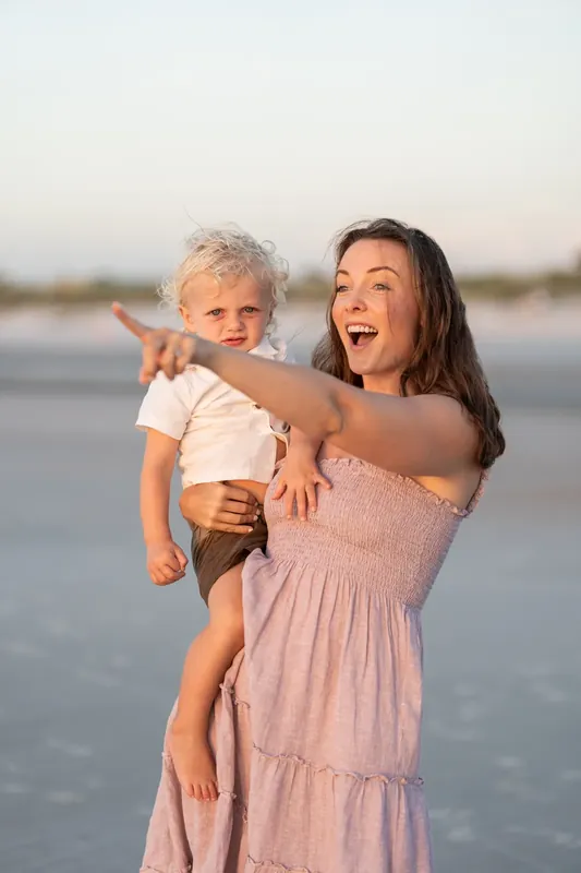 Woman in mauve tiered dress laughs while holding a blonde child on her hip at the shoreline.