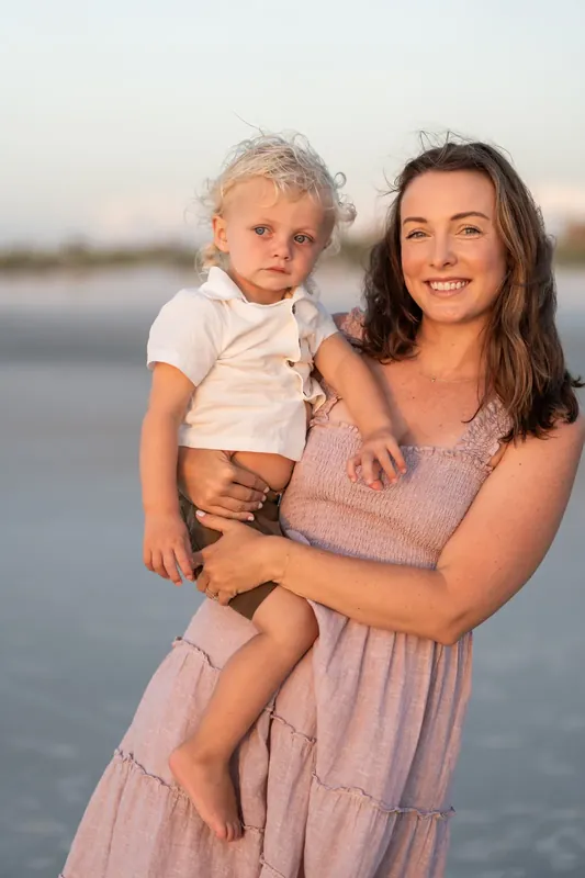 Jourdan holds a young child in a white shirt while smiling at St. Augustine Beach at sunset.