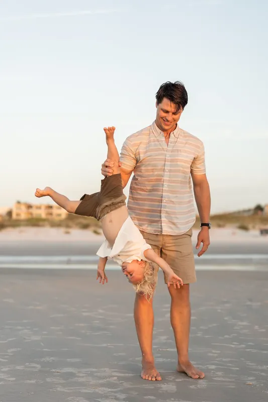 Jourdan swings their child upside down on the sand at St. Augustine Beach, both laughing.