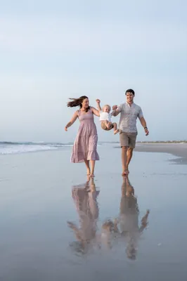 Jourdan and partner run holding toddler's hands between them, splashing through shallow water at the beach.
