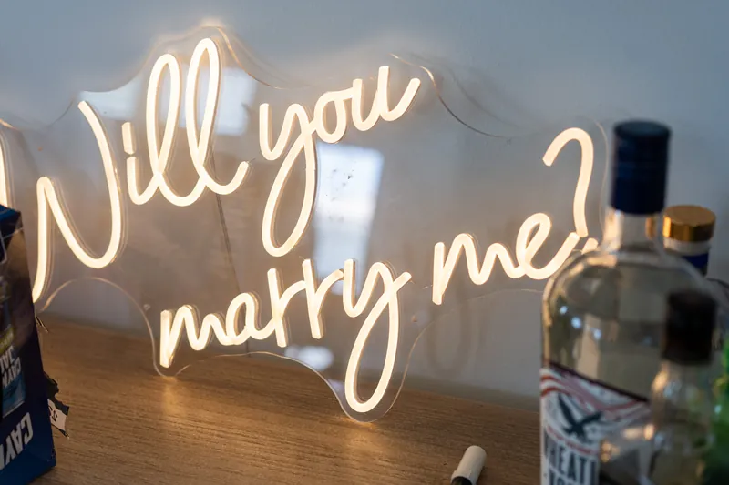 A neon sign reading "will you marry me?" glows on a table with bottles and flowers nearby.