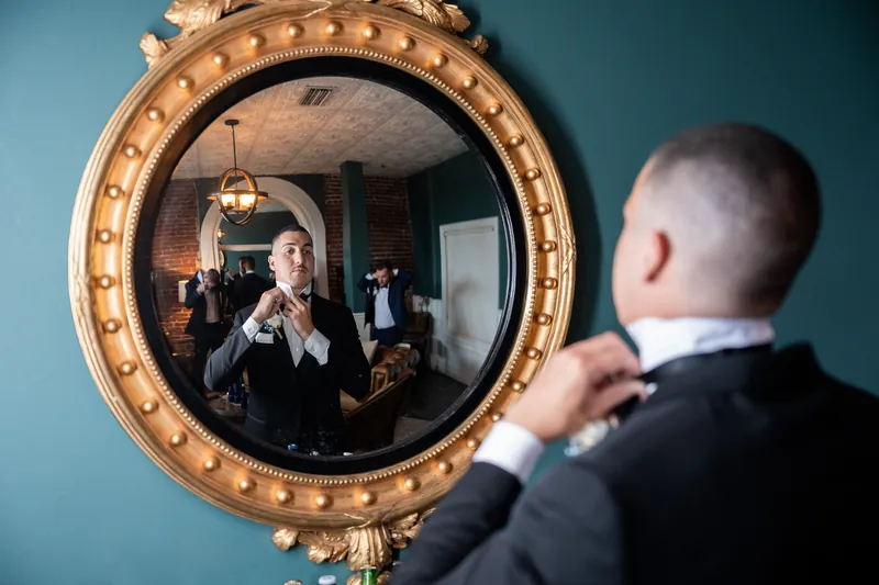Two people exchange vows reflected in round gold-trimmed mirror, one wearing white and one in dark formal attire.