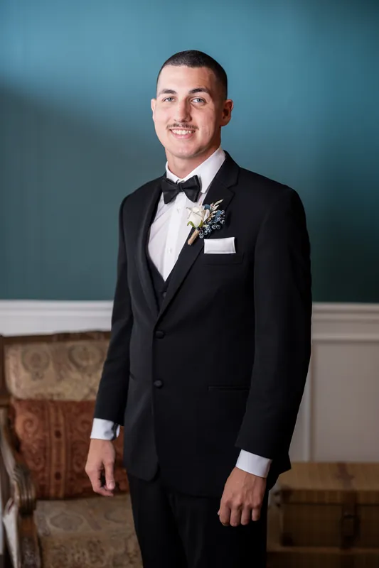 William in black tuxedo with bow tie and boutonniere stands against teal wall, smiling at camera.