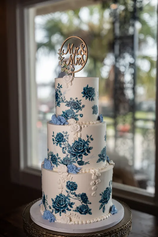Four-tier white wedding cake with blue floral design and gold monogram topper displayed near window with garden view.