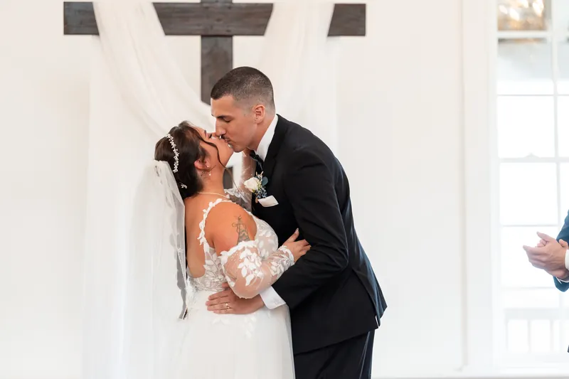 William dips Julia in a kiss at the altar, her lace gown and train flowing, with guests watching.