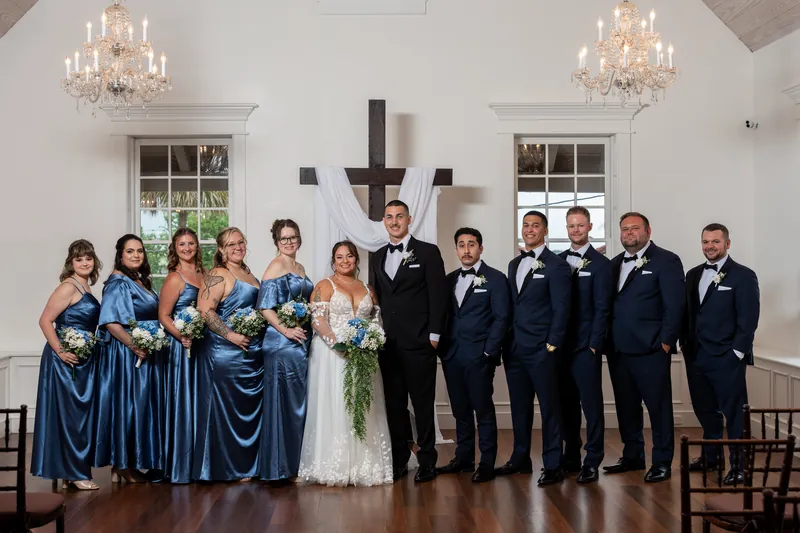 Julia in a white lace gown and William in a dark suit pose with their wedding party in navy and slate blue.
