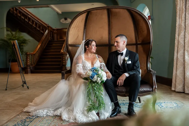 A couple sits on a leather wingback chair, holding a cascading bouquet and gazing toward each other.