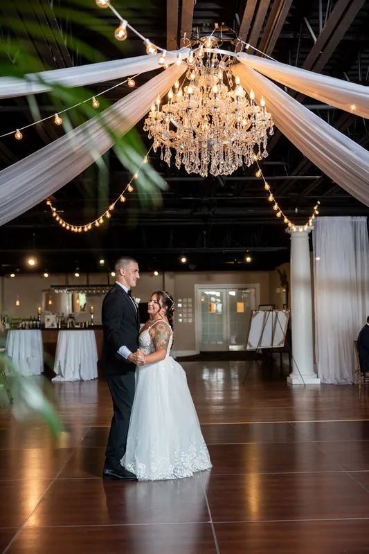 A couple dances under a crystal chandelier with draped fabric and string lights overhead.
