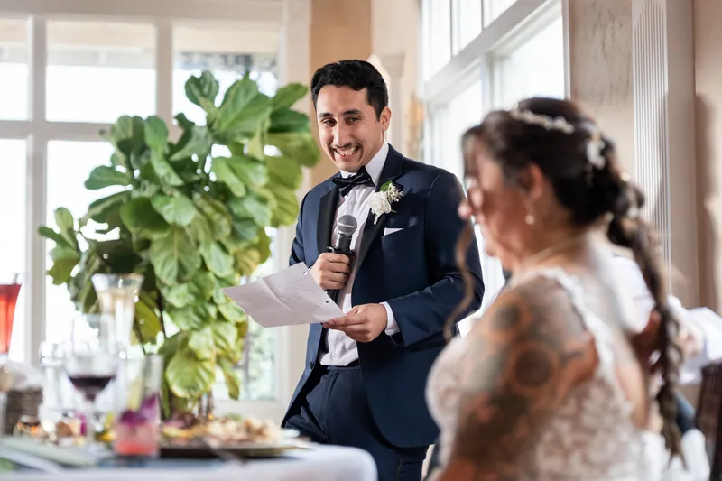 Someone in a dark suit speaks into a microphone while addressing a seated guest during the reception.