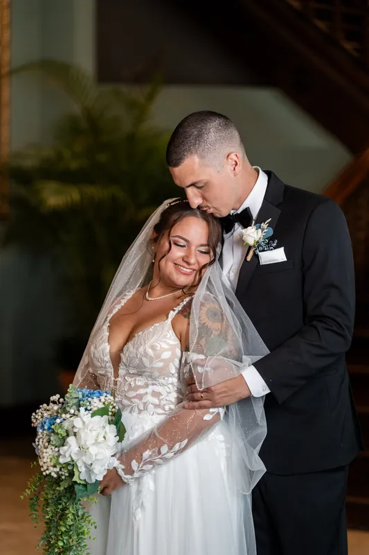 Two people embrace under soft lighting at The White Room, one in a lace gown with veil, the other in a black tuxedo.