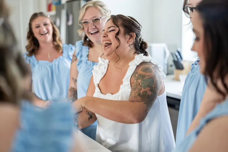 A group of people in light blue and white shirts laugh and embrace in the bridal suite during getting-ready time.