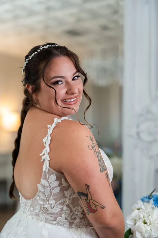 Person in white lace gown with beaded details smiles over shoulder, wearing pearl headpiece against soft interior backdrop.