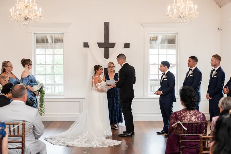 Julia and William exchange vows before guests and officiant beneath a dark cross at The White Room.