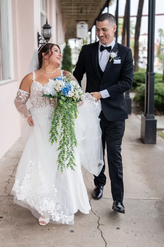 Two people walk arm-in-arm down a colonnade with white columns, both smiling as they stroll together.