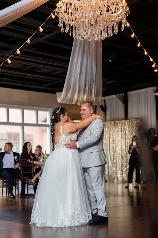 Two people share a first dance, one in white leaning into their partner as onlookers watch nearby.