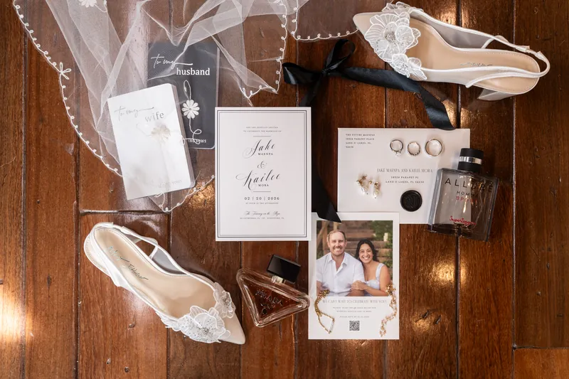 Wedding day flat lay with invitation suite, rings, shoes, perfume, and vow books on hardwood floor