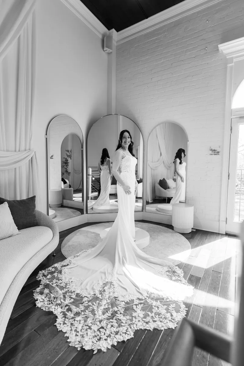 Black and white portrait of bride with lace train spread across floor in front of three arched mirrors