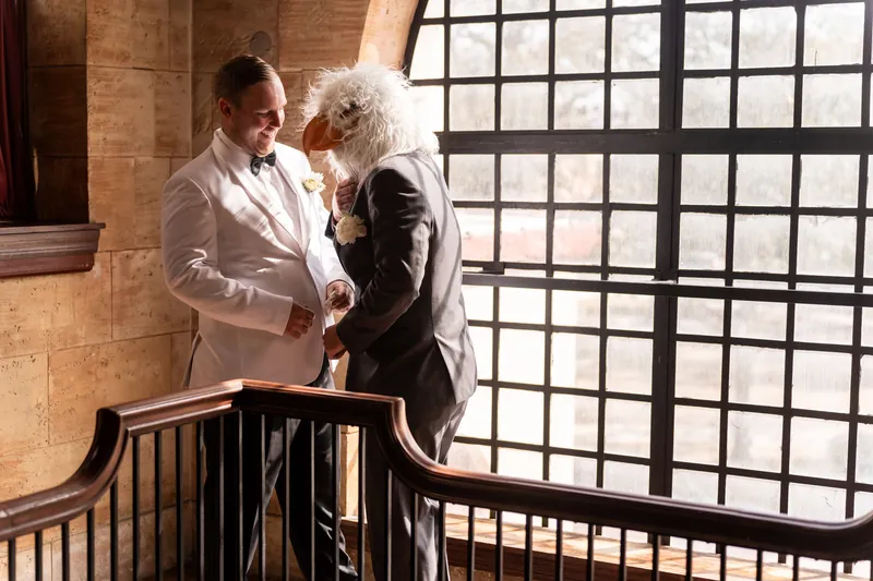 Groom laughing with groomsman in chicken mask on Treasury balcony by large windows