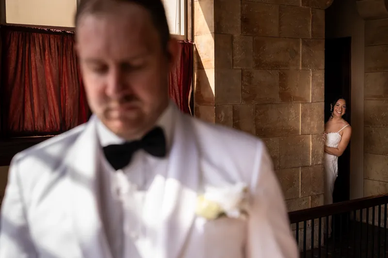 Bride peeking around stone wall smiling as groom waits for first look