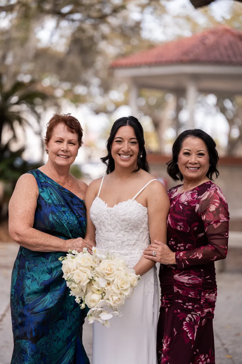Bride with both mothers holding white bouquet in the plaza with gazebo behind them