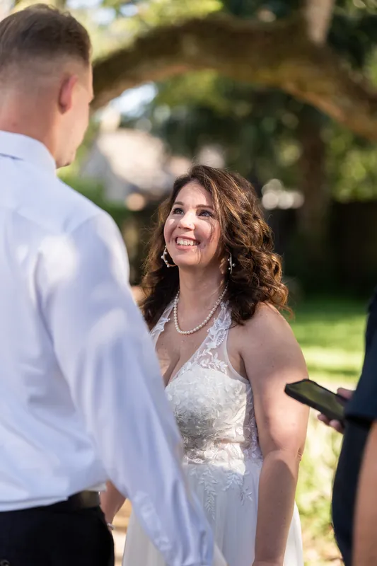 Woman in a flowing white dress with beaded bodice and pearl necklace smiles at a man in a white shirt beneath an arched garden trellis.