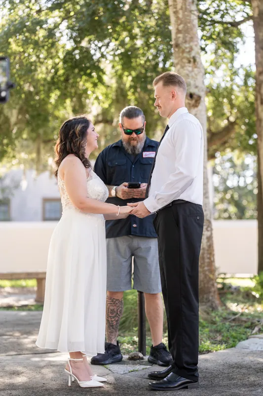 Kaley and Colton exchange vows with an officiant in a white shirt beneath a large tree in Downtown St Augustine's lush courtyard.