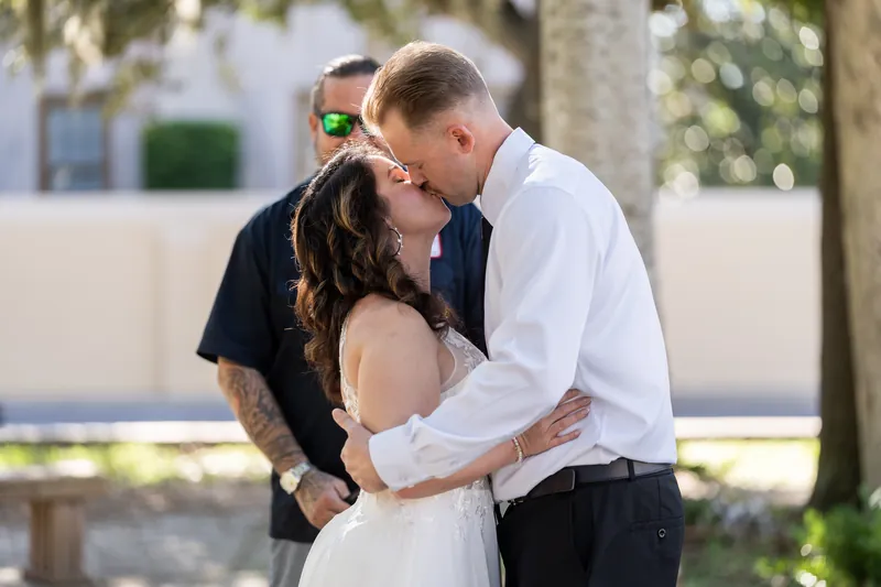 Kaley and Colton kiss under a tree while an officiant in a dark shirt watches during their ceremony in Downtown St Augustine.