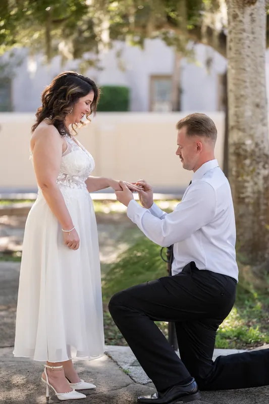 Colton kneels on one knee to slide a ring onto a woman's finger as she looks down, trees and historic architecture framing the proposal moment.