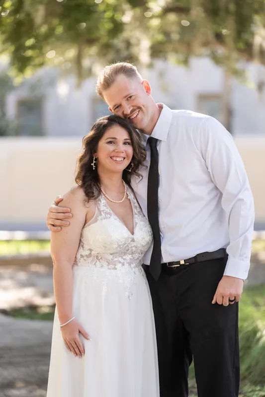 Kaley and Colton stand close together in their wedding attire beneath dappled tree shade in Downtown St Augustine, both smiling softly.