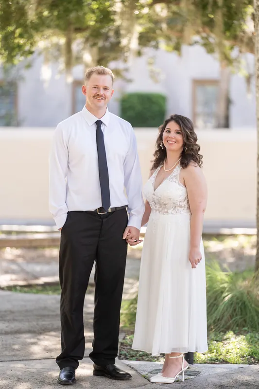 Kaley and Colton hold hands on a tree-lined street in downtown St Augustine, she in a white sleeveless dress and he in a white shirt and dark tie.