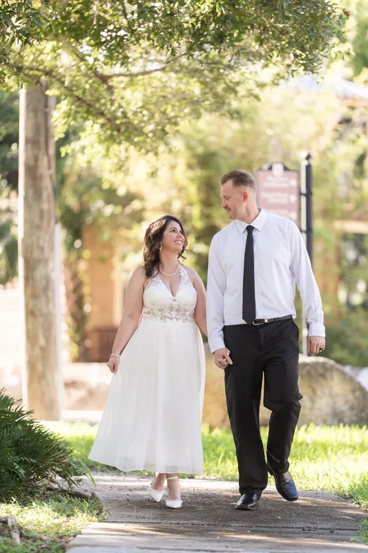 The couple walks together down a sunlit path beneath towering oaks in downtown St Augustine, her white dress flowing as they stroll hand in hand.