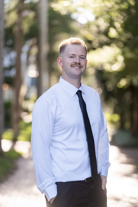A person in a white button-up shirt and dark tie stands smiling on a tree-lined street in downtown St Augustine.