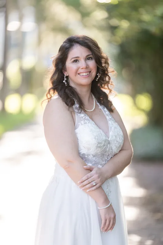 A person in a white lace gown and pearl necklace smiles while standing alone on a shaded street lined with historic trees.