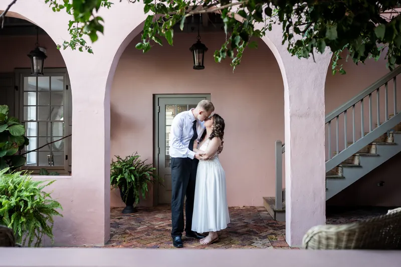 A couple in formal wedding wear kiss beneath a pink arched colonnade with ivy framing the entrance.