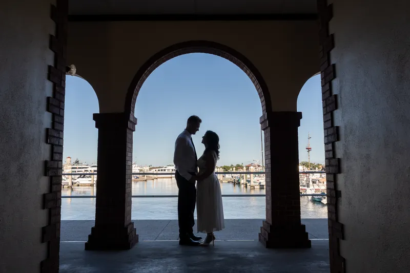 Silhouetted figures kiss in a stone archway framing a distant waterfront and clear sky beyond.