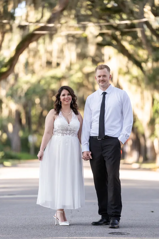 Kaley and Colton walk hand in hand down a tree-lined street in Downtown St Augustine, she in a white gown and he in a light blue shirt and dark tie.