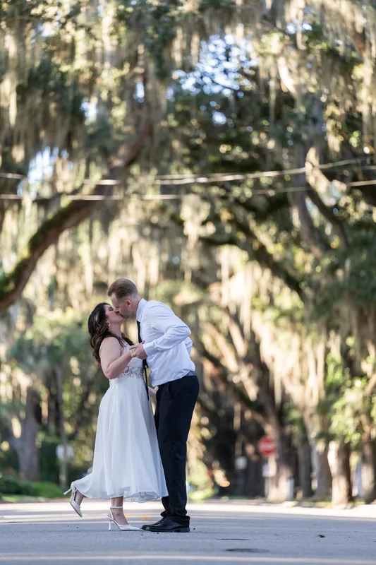 Two people kiss under Spanish moss-draped oaks on a Downtown St Augustine street, dappled light filtering through the canopy.