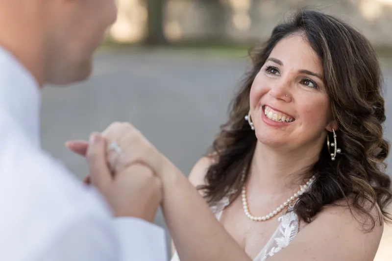 A person in a white lace gown smiles warmly while adjusting a ring on their partner's hand during an intimate moment.