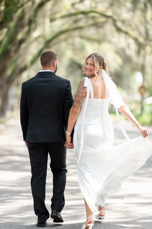 Matt and Kalsey walk hand-in-hand along a shaded tree-lined street in St. Augustine, Kalsey's white dress flowing behind