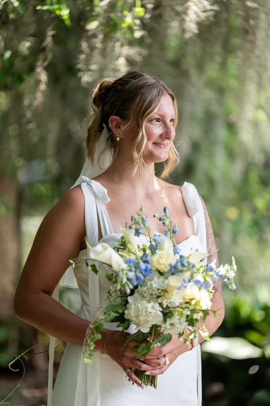 Kalsey stands beneath hanging moss holding a bouquet of white, blue, and yellow flowers, gazing thoughtfully to the side.