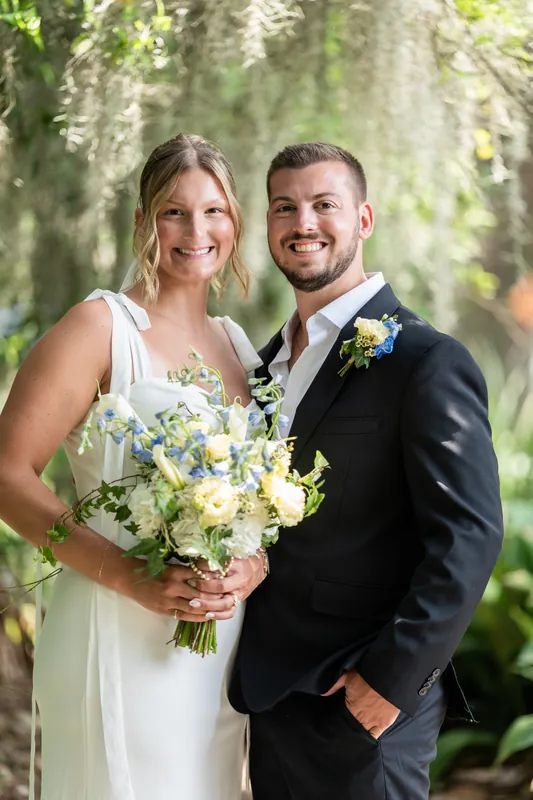 Kalsey and Matt smile together under the natural canopy of Spanish moss, both holding their white and blue bouquet.