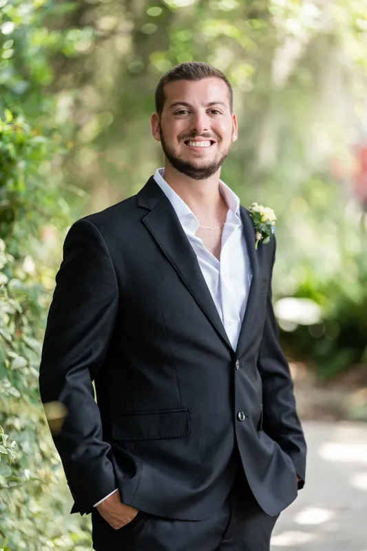 Matt stands alone under the draped trees wearing a black suit and boutonniere, smiling confidently at the camera.