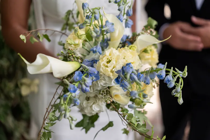 A close-up of the couple's bouquet showcasing white roses, blue delphinium, and cascading greenery with the couple blurred