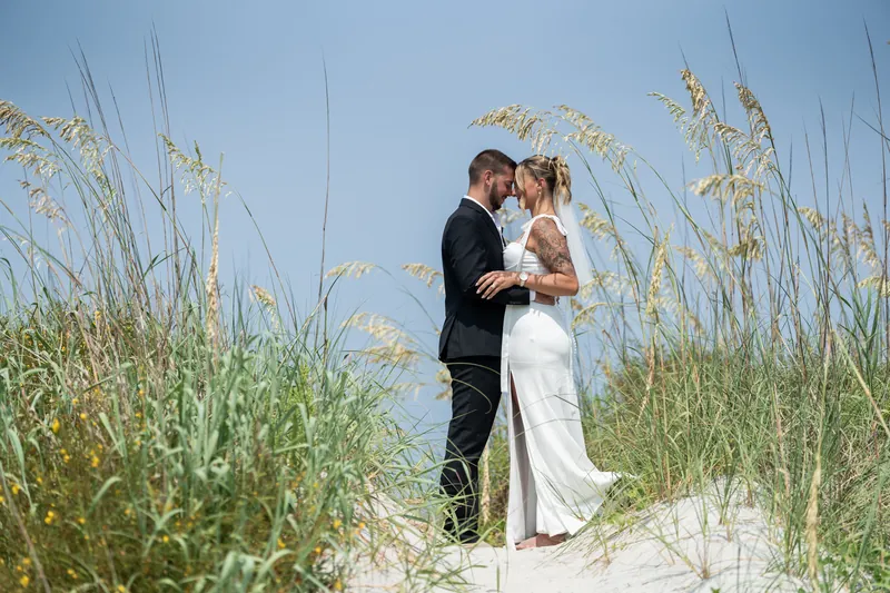Matt and Kalsey kiss among tall beach grasses, with the ocean and sky visible behind them at Vilano Beach.