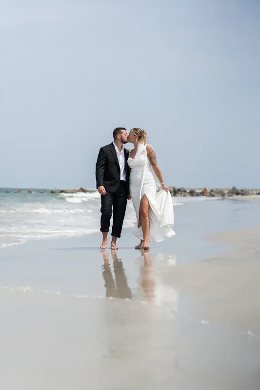 The couple walks hand in hand through shallow surf at the water's edge, their reflections visible on the wet sand.