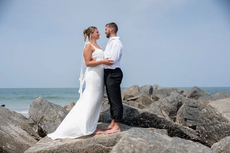 Kalsey and Matt embrace on a rocky jetty overlooking the Atlantic Ocean, their barefoot silhouettes framed against