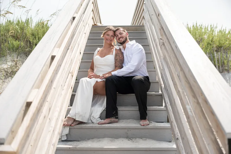 Kalsey in a white dress and Matt in a white shirt and black pants sit close together on wooden beach stairs, with dune