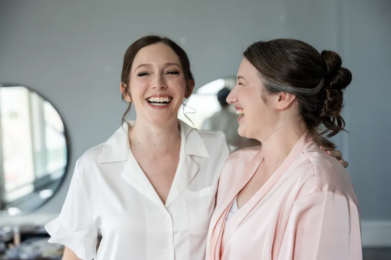 Katherine and another woman laugh together in the getting-ready suite, both wearing white and pink robes.