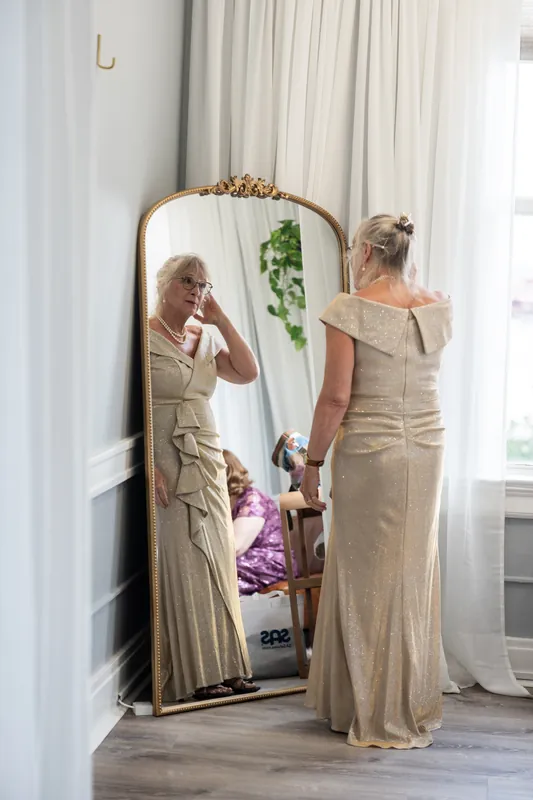 Two women in champagne-colored dresses check their appearance in a full-length mirror before the ceremony.